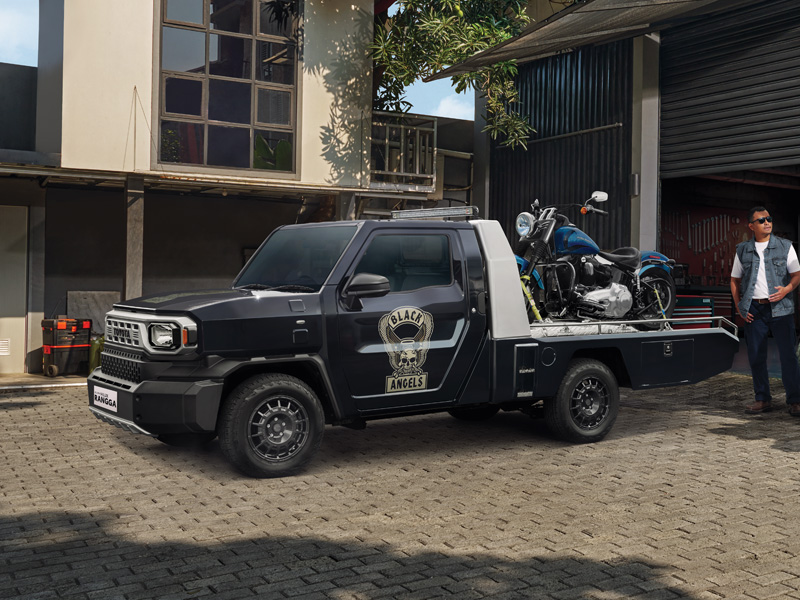 黒いHilux Rangga風のレッカー車が青と黒の大型バイクを荷台に積み、住宅街のガレージ前に停まっている。